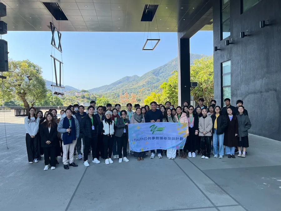 iYouth 學員與講者在戶外持計劃橫額大合照，背景為山景與玻璃幕牆。