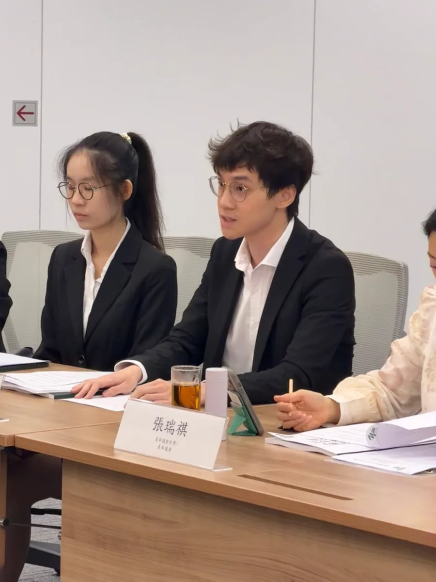 兩位青年在會議桌前交流意見，前方名牌寫有「張瑞祺」。