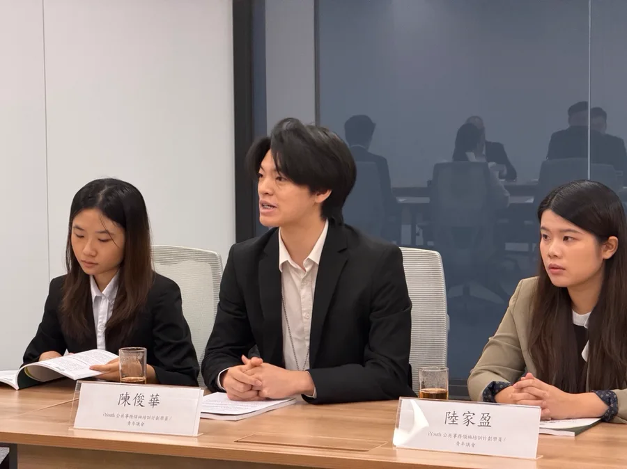 三位學員並排坐在會議桌前，中間一位正在發言，桌上有文件與座位名牌。
